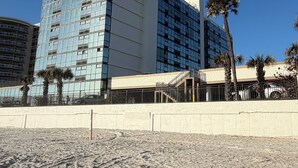 Un hôtel moderne à la façade en verre, avec une plage de sable en premier plan et des palmiers.