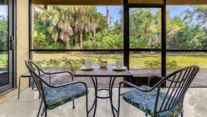 A covered patio with two chairs and a table, large windows offering a view of a garden with palm trees and other greenery. - Tranquil Sarasota Retreat Near Beaches, Pool & Nature Escape - Timberwood Villa #183 - Roelens (Sarasota)