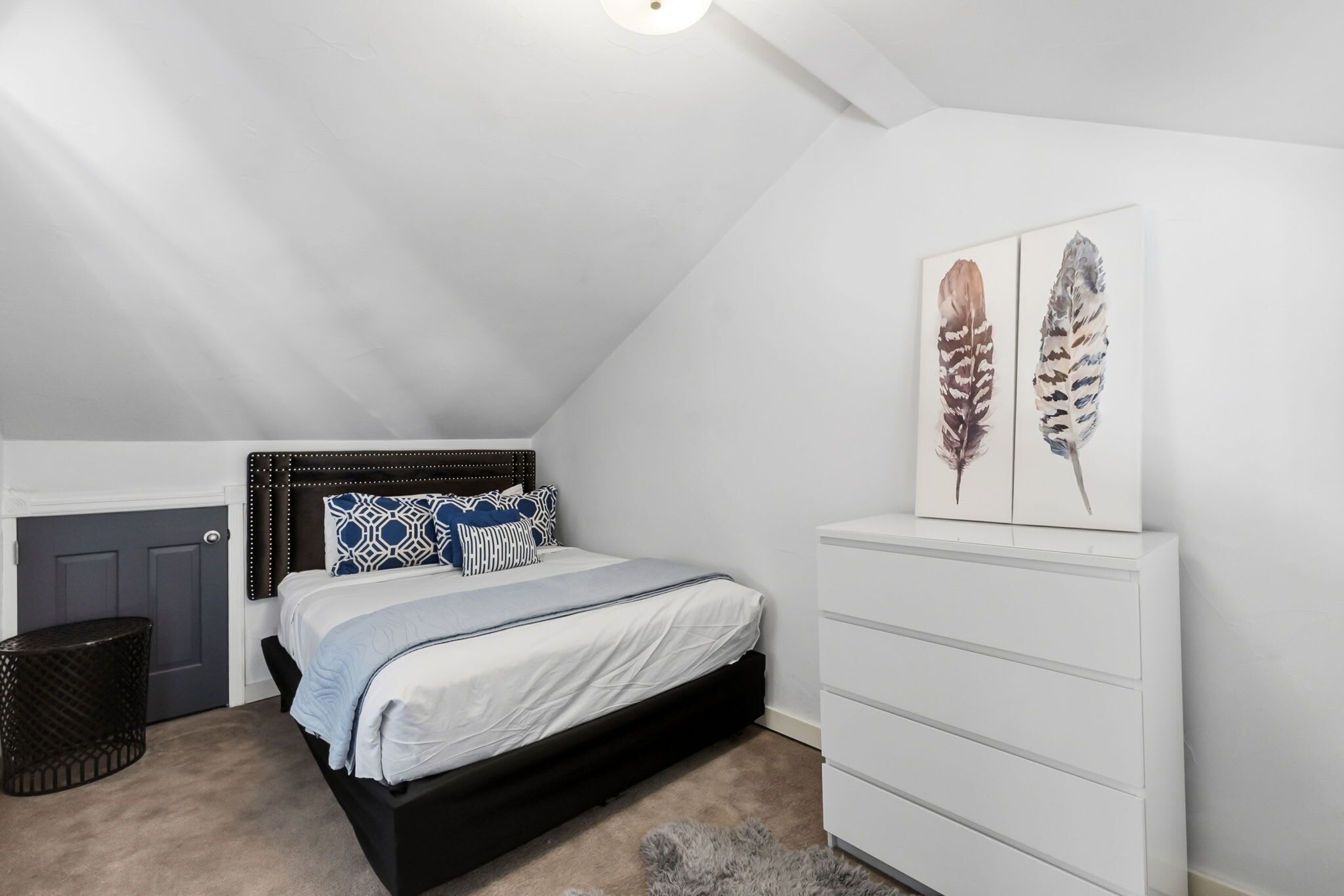 A bedroom with a sloped ceiling, a bed with a black frame, a white dresser, and two feather wall art pieces.