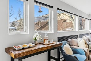 A room with a wooden table, a blue chair, and a view of a brick building through large windows. - East Vail Escape - Mountain Views & Shared Hot Tub (Vail)