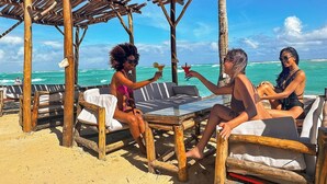 Un área de descanso frente a la playa con muebles de madera y una mesa de vidrio, con vista al océano.