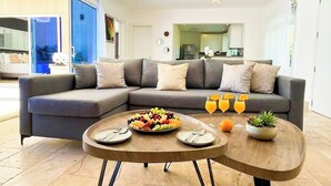 Una sala de estar moderna con un sofá seccional gris, una mesa de centro de madera y una mesa de comedor dispuesta con platos, cubiertos y una bandeja de fruta.