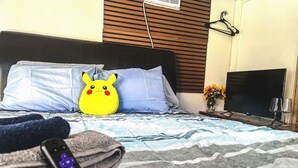Un lit avec une couverture à rayures, un oreiller orné d’un motif Pikachu, une télécommande, un téléviseur et un vase rempli de fleurs.
