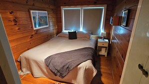 A wooden cabin bedroom with a bed, nightstand, and a window with blinds. - Beachfront Property in Private Bay with only 12 houses and private beach access. (Matapouri)