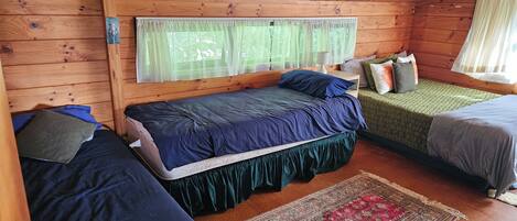 A cabin with two beds, a wooden ceiling, a window with sheer curtains, and a small table with a lamp.