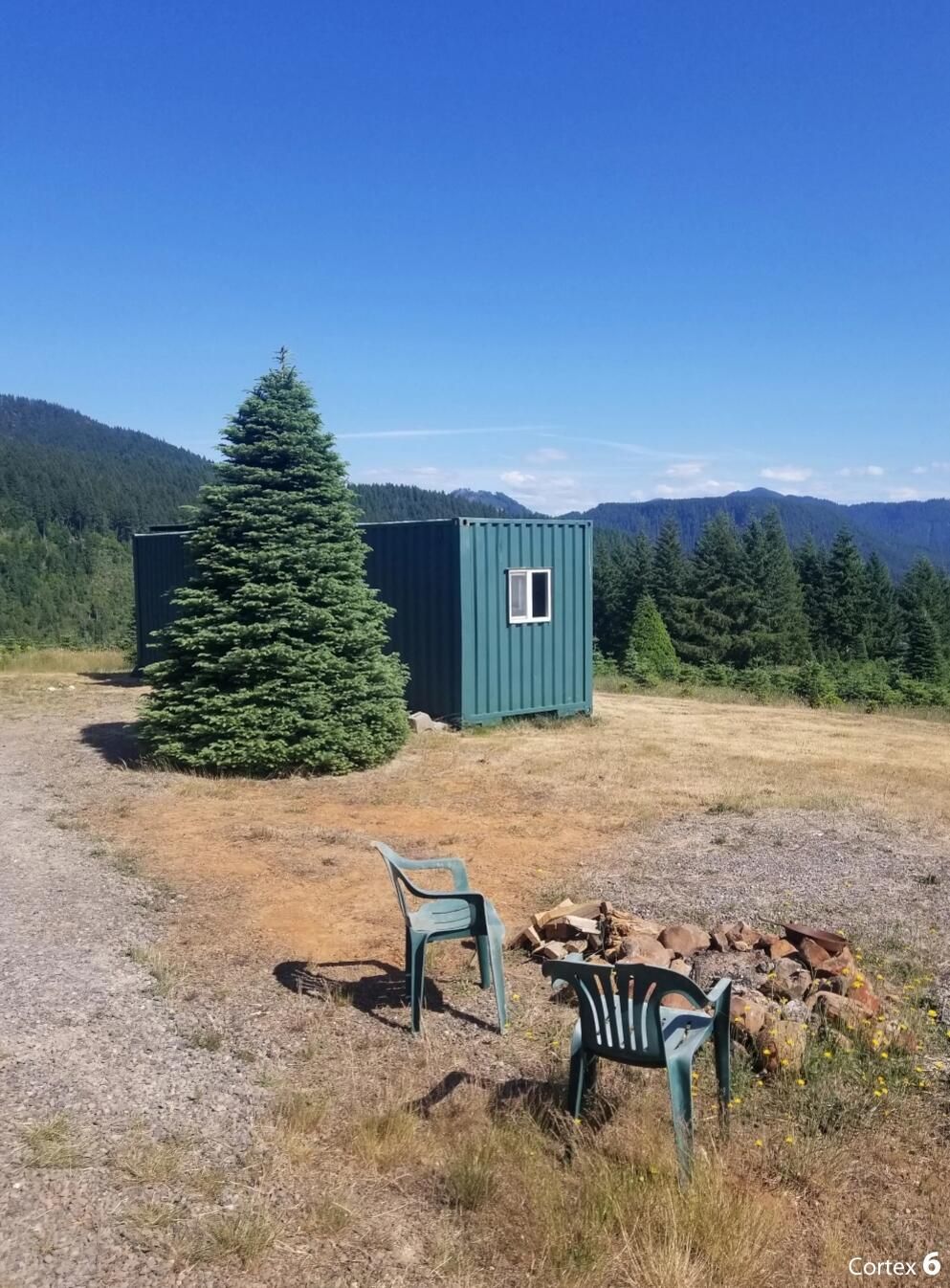 A small green cabin with a window, surrounded by a forest and mountains.
