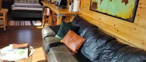 A cozy cabin interior with a leather sofa, a bed with plaid bedding, a wooden dining table, and a world map on the wall.