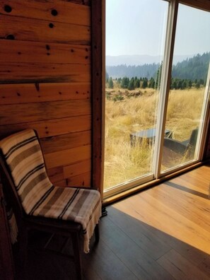 A wooden cabin interior with a large window offering a view of a grassy field and distant trees. - Hot tub! sought out Christmas tree heaven experience. unique container. (Oakridge)