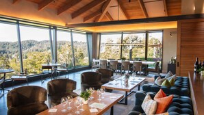 A spacious dining area with large windows offering a view of the forest. Tables are set with wine glasses and plates. - Hummingway Ranch, by Vinifera Homes (Forestville)