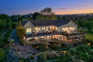 Une maison à deux étages dotée d’une terrasse et d’un espace salon extérieur, le tout entouré d’un jardin aménagé.