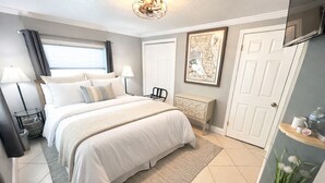 Un dormitorio con cama, lámparas de noche, un radiador, una puerta, un estante en la pared y una ventana con cortinas.