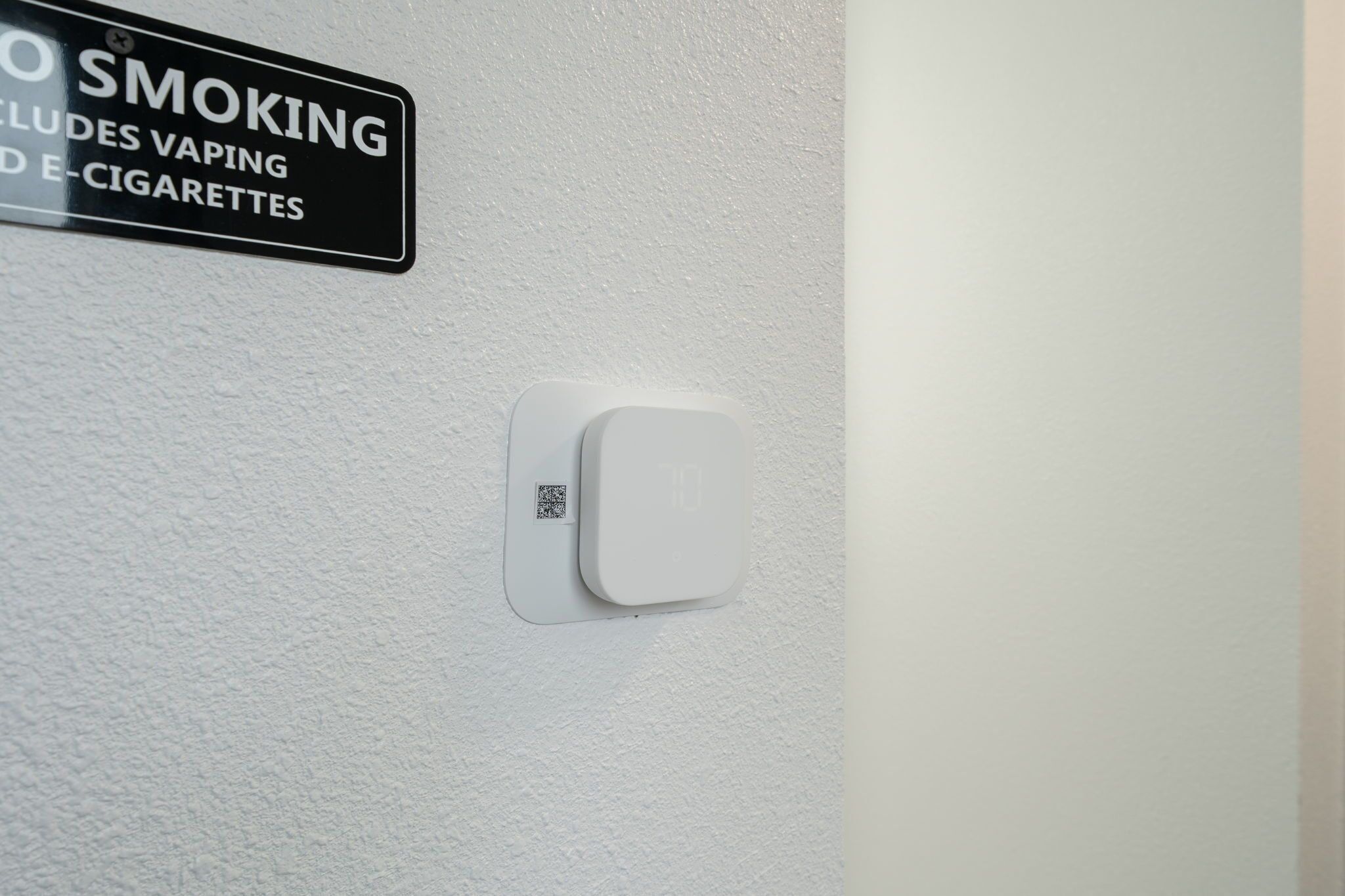 Señalización de prohibido fumar, incluyendo vapeo y cigarrillos electrónicos, con un código QR y un detector de humo en una pared blanca.