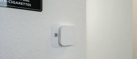 Señalización de prohibido fumar, incluyendo vapeo y cigarrillos electrónicos, con un código QR y un detector de humo en una pared blanca.