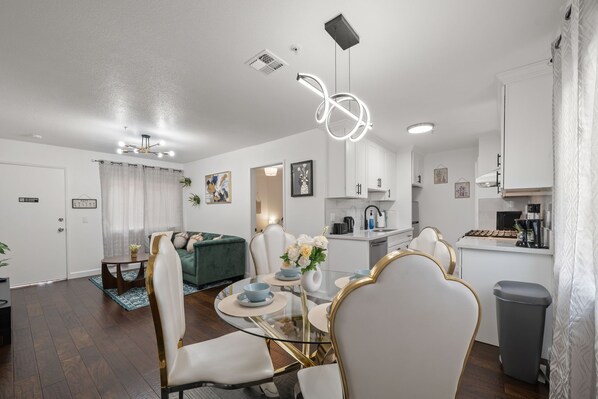 Una sala de estar moderna con una mesa de comedor de vidrio, sillas blancas, un sofá verde y una zona de cocina con armarios blancos y una cafetera.