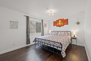 Un dormitorio con cama, mesita de noche, lámpara de pared, ventana con cortinas y una obra de arte en la pared.