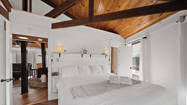 Ein geräumiges Schlafzimmer mit einem großen Bett, Holzbalken an der Decke und Blick durch eine offene Tür in einen Wohnbereich.