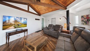 Ein geräumiges Wohnzimmer mit Holzboden, einem großen Flachbildfernseher an der Wand, einem Ledersofa und einem Couchtisch aus Holz.