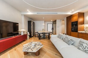 A modern living room with a flat-screen TV, a red entertainment unit, a marble coffee table, a white sofa with patterned cushions, and a dining area with a round table and chairs. - Lakeside Luxe Apartment 2BR Prime Location (Tirana)