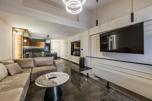 Un salon moderne avec un canapé d’angle, une table basse ronde, un téléviseur à écran plat fixé au mur et un coin cuisine visible à travers une porte ouverte.