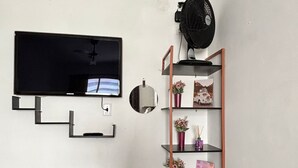 Een kamer met een flatscreen televisie aan de muur, een kleine tafel met een witte tafelkleed, een hoge houten plank met decoratieve objecten, en een zwarte ventilator aan de muur.