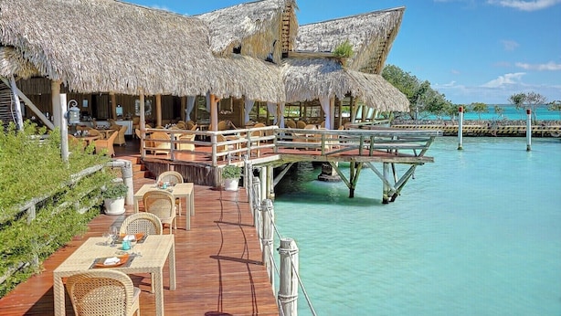 Bungalows sobre el agua con techos de paja, asientos en terraza de madera y vista al mar turquesa.