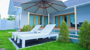 Une maison moderne avec une piscine, des chaises longues blanches et un grand parasol.