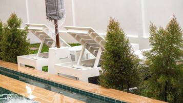 Un área junto a la piscina con sillas reclinables blancas y un pequeño árbol.