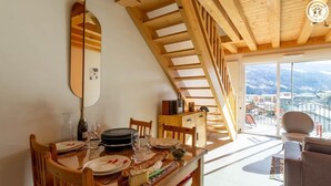 Un comedor con una mesa de madera dispuesta para dos personas, una escalera de madera y vistas a un pueblo a través de amplios ventanales.
