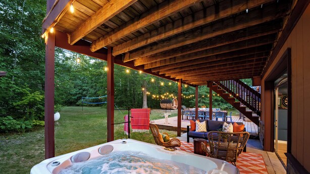 Un jacuzzi bajo una pérgola de madera con luces colgantes, rodeado de mobiliario exterior y una escalera que conduce a un nivel superior.