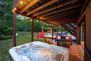 Un jacuzzi bajo una pérgola de madera con luces colgantes, rodeado de mobiliario exterior y una escalera que conduce a un nivel superior.