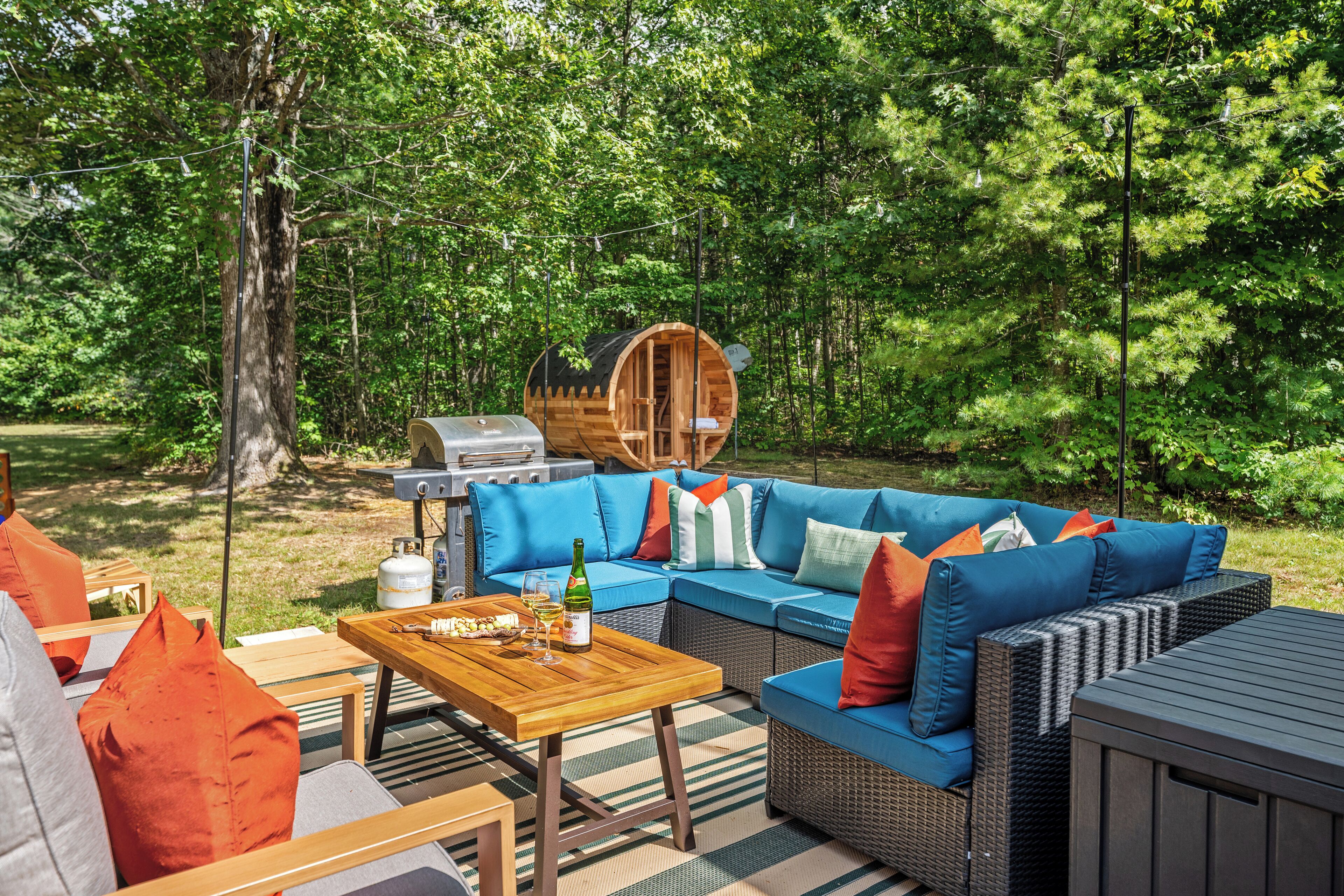 A wooden deck with a blue sofa, orange pillows, and a wooden table with a bottle and glasses.