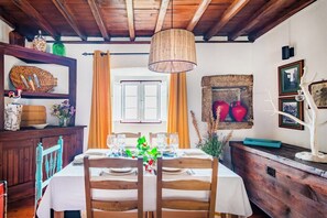 A dining area with a table set for four, wooden chairs, a hanging light fixture, and a stone wall with decorative items. - Casa das 10 Janelas at Monsanto (Monsanto)