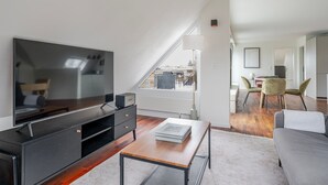 Ein modernes Wohnzimmer mit Flachbildfernseher, Couchtisch und Sofa. Im Hintergrund ist ein Essbereich mit Stühlen und Tisch zu sehen.