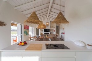 Eine moderne Küche mit weißer Arbeitsplatte, zwei hängenden Rattan-Leuchten, einem Essbereich mit Tisch und Stühlen sowie Blick ins Freie.