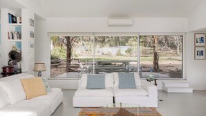Una sala de estar moderna con un sofá blanco, una mesa de centro de cristal y ventanas amplias que dan a un jardín con mobiliario exterior.