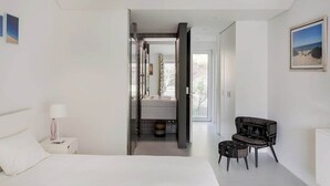 Un dormitorio con cama, mesita de noche, una silla y un baño visible a través de una puerta abierta.