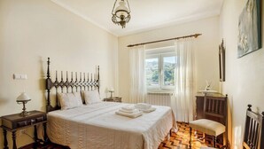 Een slaapkamer met een groot bed, een houten vloer, een raam met gordijnen, een nachtkastje met een lamp en een stoel.