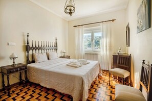 Ein Schlafzimmer mit einem großen Bett, Holzboden, einem Fenster mit Vorhängen, einem Nachttisch mit Lampe und einem Sessel.