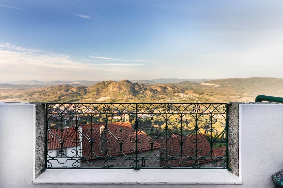 A balcony with a wrought iron railing overlooking a mountainous landscape with red-roofed buildings.
