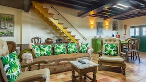 A spacious living area with a wooden staircase, a sofa with tropical cushions, a chair with a cushion, and a small table with a remote control.