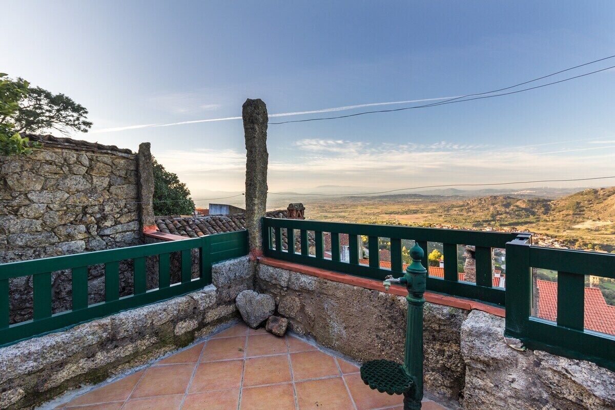 Un muro de piedra con barandilla verde, suelo de baldosas y vista a colinas distantes.