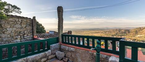 Un muro de piedra con barandilla verde, suelo de baldosas y vista a colinas distantes.