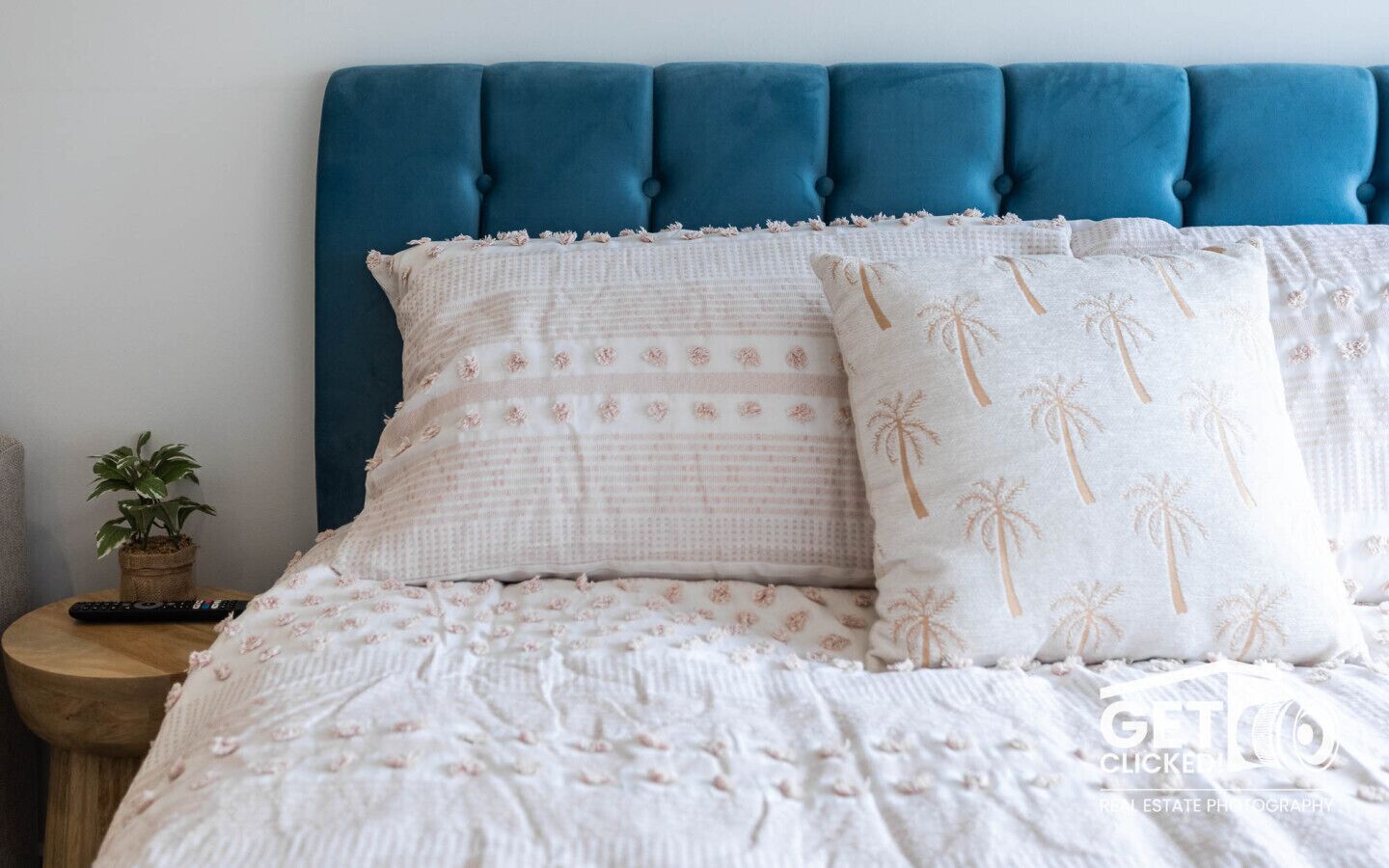 A bed with a blue upholstered headboard, a white bedspread with a palm tree pattern, and a small potted plant on a wooden nightstand.
