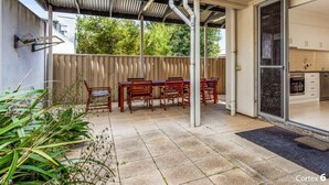 Eine überdachte Terrasse mit Esstisch und Stühlen, eine Schiebetür zu einer Küche und eine Dachkonstruktion aus Metall.