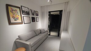 A modern living room with a grey sofa, a brown stool, and a dining table. There are framed pictures on the wall and a sliding door leading to another room. - 1-Bedroom Apartment - Comfort and relaxation with complete facilities and easy access to Carnival. (Salvador)