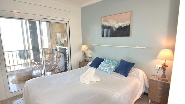 Slaapkamer met een bed, twee nachtkastjes, een wandlamp, een schilderij en een schuifdeur naar een balkon met rieten meubilair.