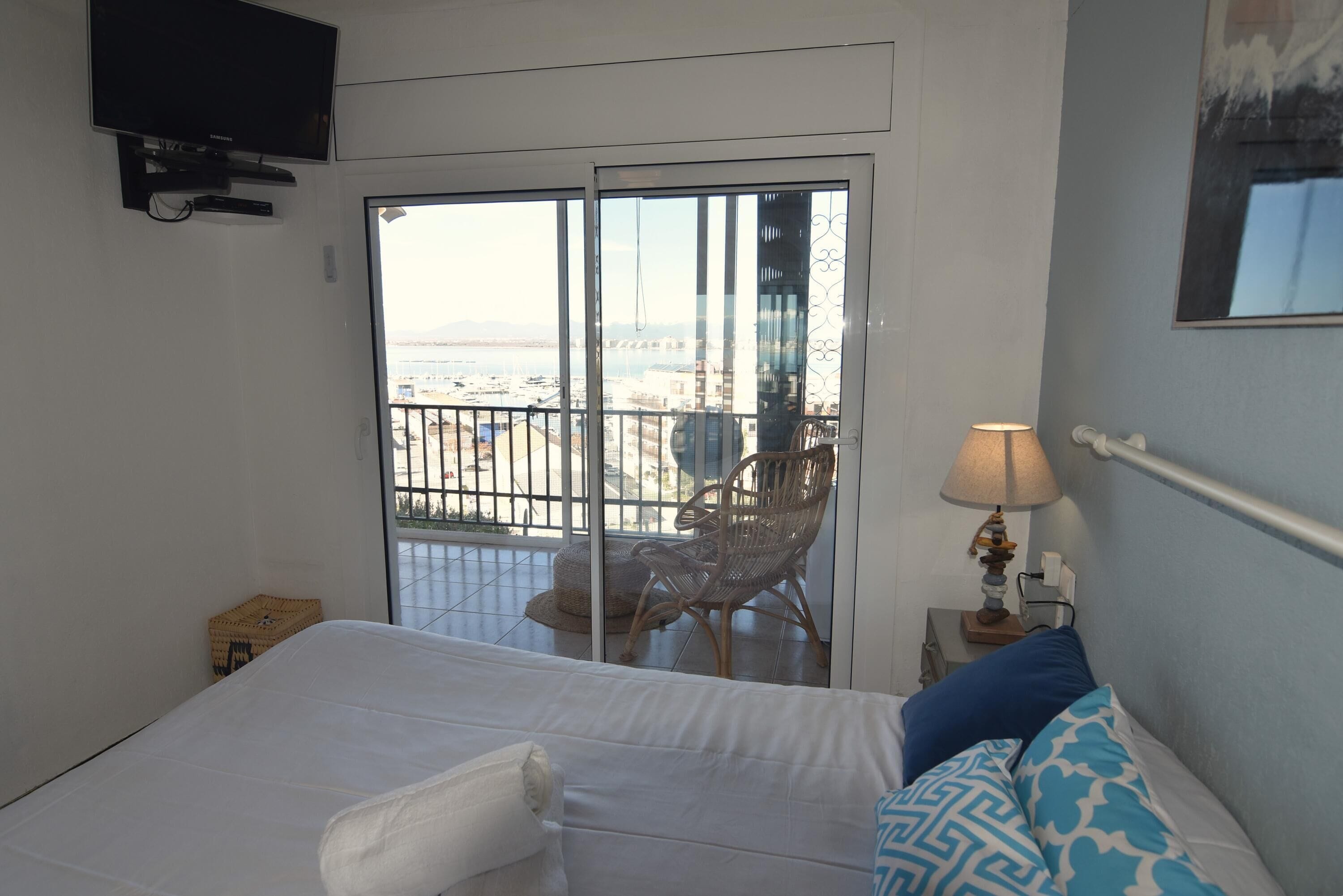 A bedroom with a bed, a TV mounted on the wall, a balcony with a chair, and a lamp on a bedside table.