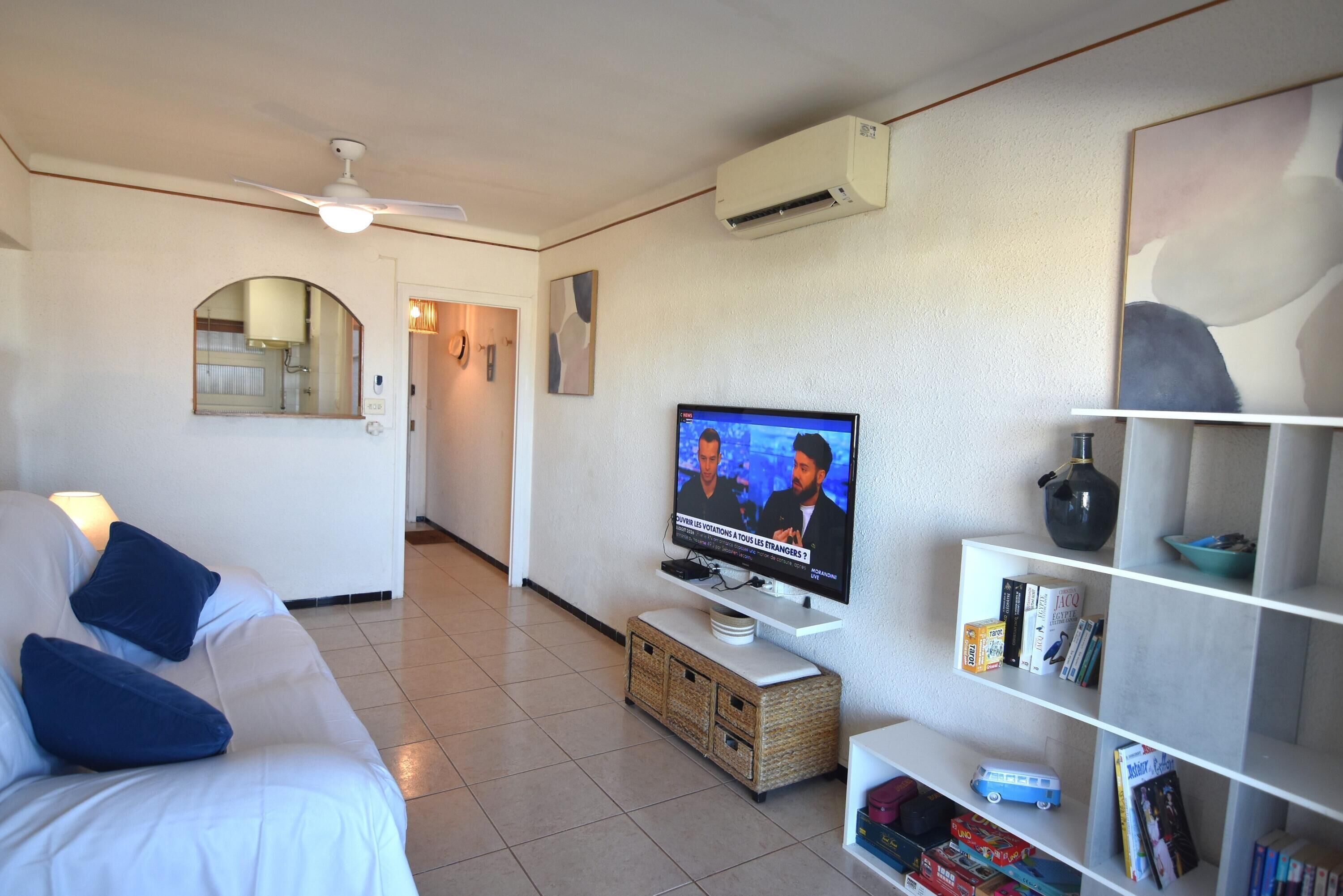 Una sala de estar moderna con un televisor de pantalla plana, un sofá blanco con cojines azules, una mesita de centro, una estantería y un ventilador de techo.