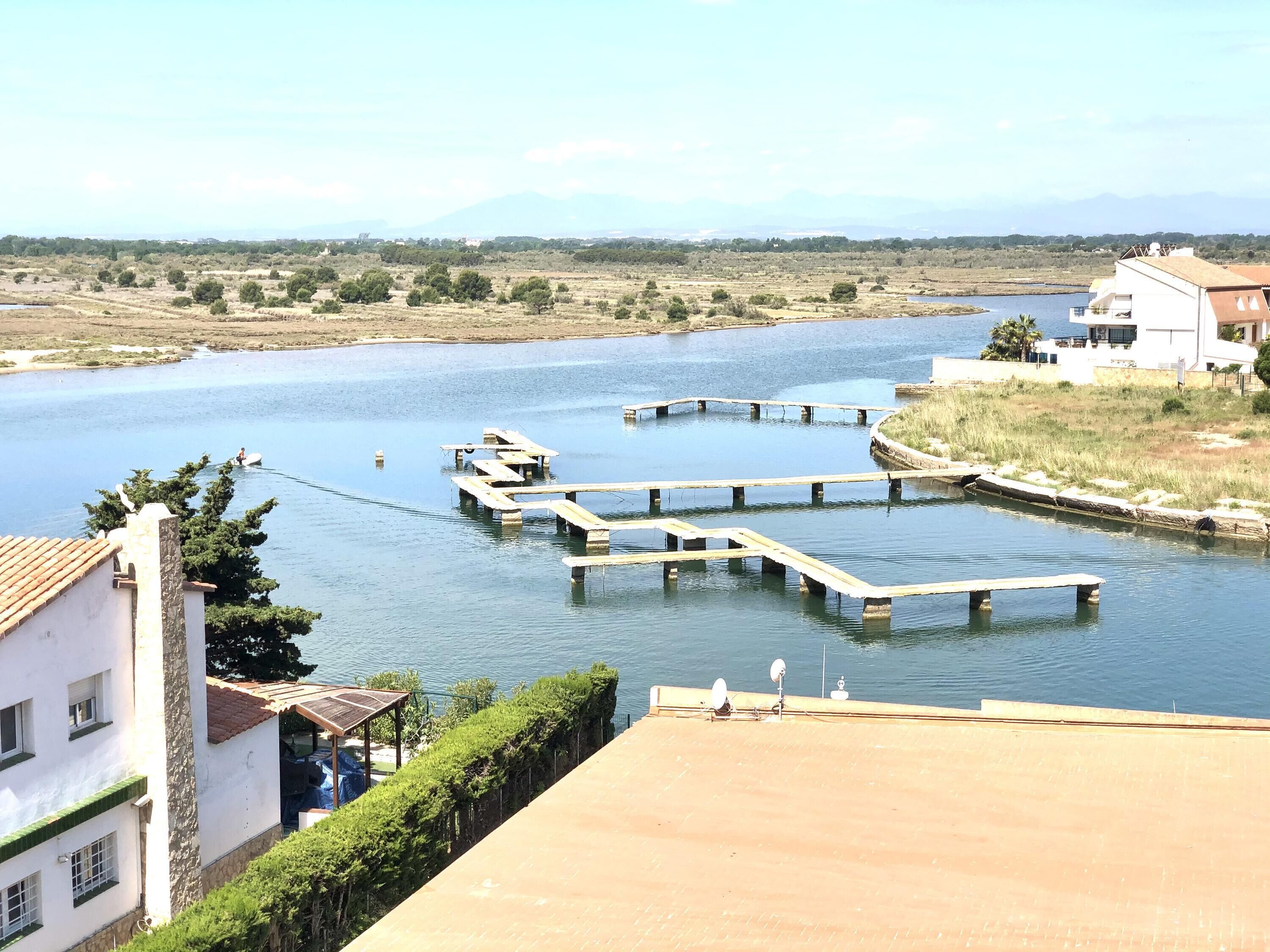 Uitzicht op een rivier met een houten steiger die in het water reikt, omgeven door begroeiing en gebouwen.
