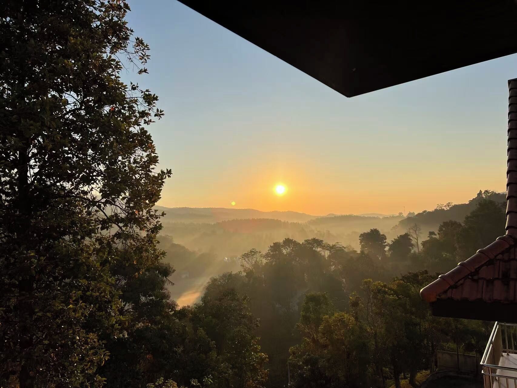 A sunrise over a hilly landscape with trees and a building in the foreground.